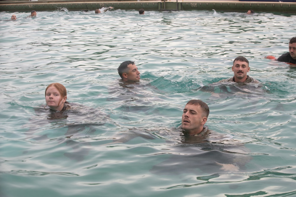 Multiple individuals in military-style uniforms or clothing are swimming in an indoor pool, suggesting a military water survival or swim qualification training exercise. At least four people are clearly visible in the foreground, with additional swimmers visible in the background. The participants appear to be treading water while fully clothed, which is consistent with combat water survival training.