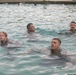 Multiple individuals in military-style uniforms or clothing are swimming in an indoor pool, suggesting a military water survival or swim qualification training exercise. At least four people are clearly visible in the foreground, with additional swimmers visible in the background. The participants appear to be treading water while fully clothed, which is consistent with combat water survival training.