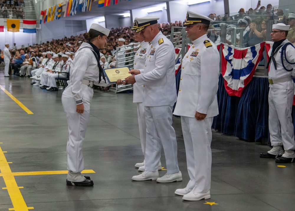 A U.S. Navy graduation ceremony is taking place in a large indoor facility, likely a recruit training command. A junior enlisted sailor is receiving a certificate or award from a senior officer in dress white uniforms. A large audience of sailors and civilians fills the bleachers in the background.