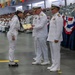 A U.S. Navy graduation ceremony is taking place in a large indoor facility, likely a recruit training command. A junior enlisted sailor is receiving a certificate or award from a senior officer in dress white uniforms. A large audience of sailors and civilians fills the bleachers in the background.