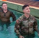 Three military personnel are standing in a pool wearing their full uniforms, appearing to participate in a water survival or combat swimming training exercise. The foreground figure is a U.S. Army soldier identified by name tape as 'Benedict,' wearing OCP (Operational Camouflage Pattern) uniform with a U.S. flag patch and what appears to be a rank insignia. Behind him are two individuals in U.S. Marine Corps MARPAT (digital camouflage) uniforms.