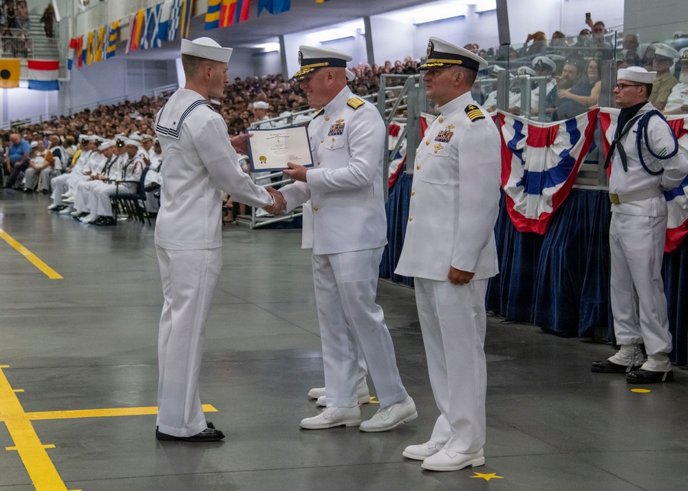 A U.S. Navy graduation or awards ceremony is taking place in a large indoor facility. A senior officer in white dress uniform is presenting a certificate or award to an enlisted sailor while another officer stands nearby. The venue is filled with seated sailors and civilian spectators in the background.