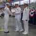 A U.S. Navy graduation or awards ceremony is taking place in a large indoor facility. A senior officer in white dress uniform is presenting a certificate or award to an enlisted sailor while another officer stands nearby. The venue is filled with seated sailors and civilian spectators in the background.