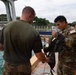 Military personnel are gathered near a waterfront facility, with a U.S. Army soldier wearing multicam uniform (unit number 515 visible) handling what appears to be a tactical vest or harness. An instructor or fellow service member in an olive green t-shirt and multicam pants is assisting or demonstrating equipment handling. A third service member is visible in the background, suggesting a training or equipment inspection scenario.