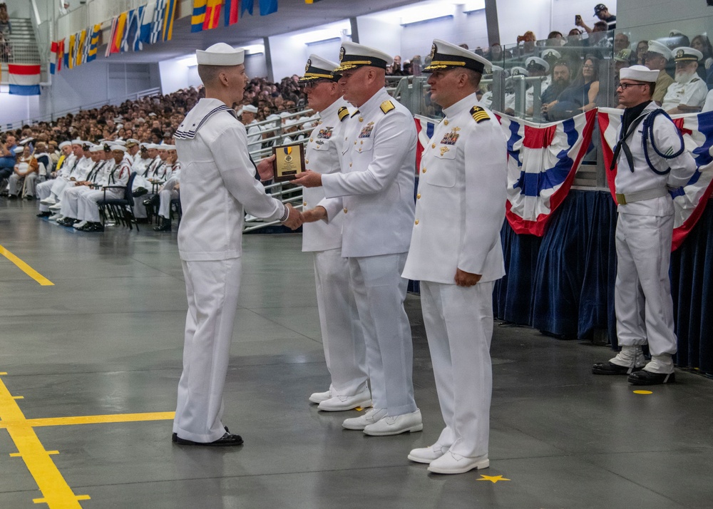 A formal U.S. Navy military ceremony is taking place in a large indoor venue, likely a graduation or awards ceremony. A junior enlisted sailor in dress whites is receiving a plaque or award from senior officers, one of whom appears to hold the rank of Admiral based on gold sleeve stripes. A large audience of uniformed personnel and civilians fills the background.