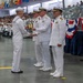 A formal U.S. Navy military ceremony is taking place in a large indoor venue, likely a graduation or awards ceremony. A junior enlisted sailor in dress whites is receiving a plaque or award from senior officers, one of whom appears to hold the rank of Admiral based on gold sleeve stripes. A large audience of uniformed personnel and civilians fills the background.