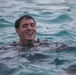 A young man is swimming in open water, with his head and upper shoulders visible above the surface. He is smiling broadly with his eyes closed, expressing joy or exhilaration. He appears to be wearing a dark wetsuit or dark clothing.