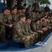 A large group of military personnel in camouflage uniforms are seated in bleachers or stadium-style seating, appearing to attend a briefing or event. The soldiers appear wet, suggesting recent water-based training activity. The setting appears to be an outdoor facility in a tropical environment, indicated by palm trees visible in the background.