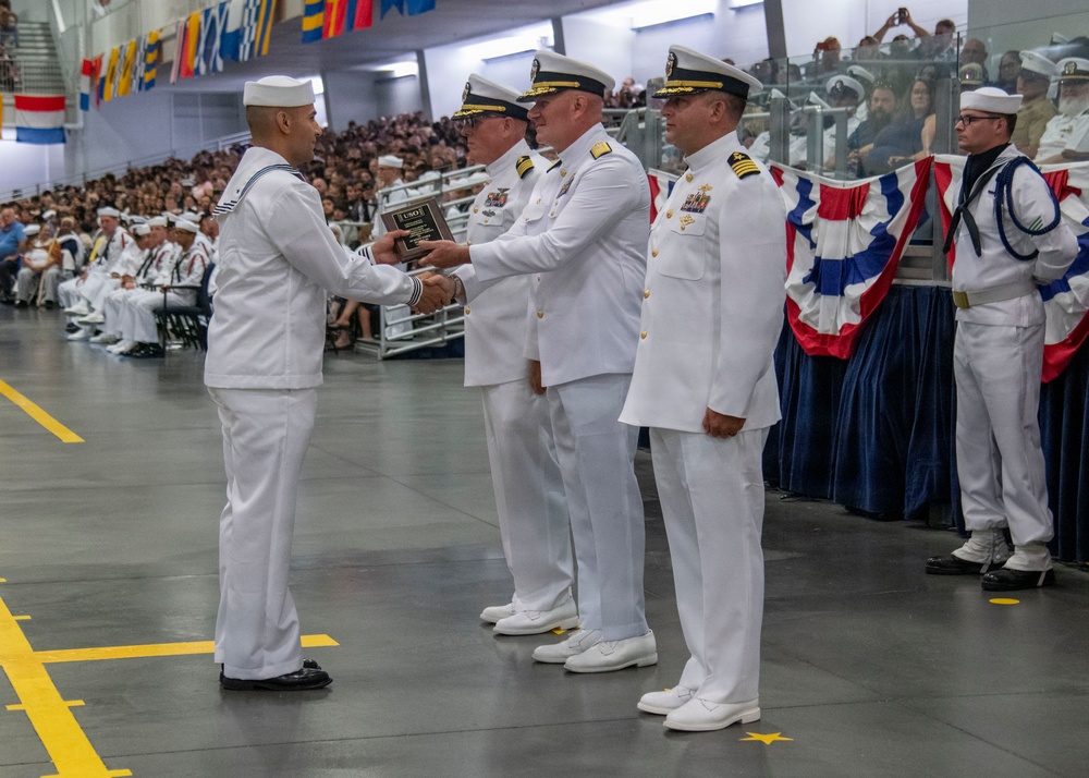 A formal U.S. Navy military ceremony is taking place in a large indoor venue, where a junior enlisted sailor is receiving what appears to be a USO award plaque from a senior officer. Multiple officers in white dress uniforms are present on the floor, while a large audience of sailors and civilians fills the bleachers in the background. The venue is decorated with nautical signal flags and red, white, and blue bunting.
