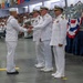 A formal U.S. Navy military ceremony is taking place in a large indoor venue, where a junior enlisted sailor is receiving what appears to be a USO award plaque from a senior officer. Multiple officers in white dress uniforms are present on the floor, while a large audience of sailors and civilians fills the bleachers in the background. The venue is decorated with nautical signal flags and red, white, and blue bunting.