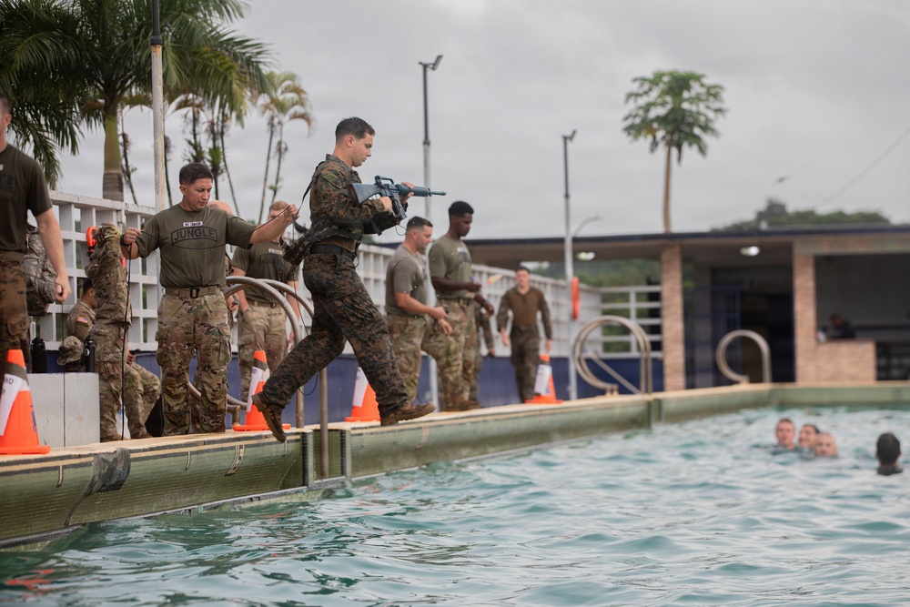 Military personnel are conducting a water survival or combat swimming training exercise at an outdoor pool facility. A Marine in woodland MARPAT camouflage is jumping into the pool while carrying a rifle, while other service members in various uniforms observe or wait their turn along the pool deck. Several swimmers are already visible in the water, and orange traffic cones mark the training area.
