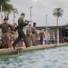 Military personnel are conducting a water survival or combat swimming training exercise at an outdoor pool facility. A Marine in woodland MARPAT camouflage is jumping into the pool while carrying a rifle, while other service members in various uniforms observe or wait their turn along the pool deck. Several swimmers are already visible in the water, and orange traffic cones mark the training area.