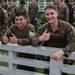 Two military personnel in camouflage uniforms lean against a white painted fence, both giving thumbs-up gestures and smiling at the camera. They appear to be at a military training event or exercise, with numerous other uniformed personnel visible in the background. One individual has a numbered tag 'W116' visible on their sleeve, suggesting a competition or evaluation event.