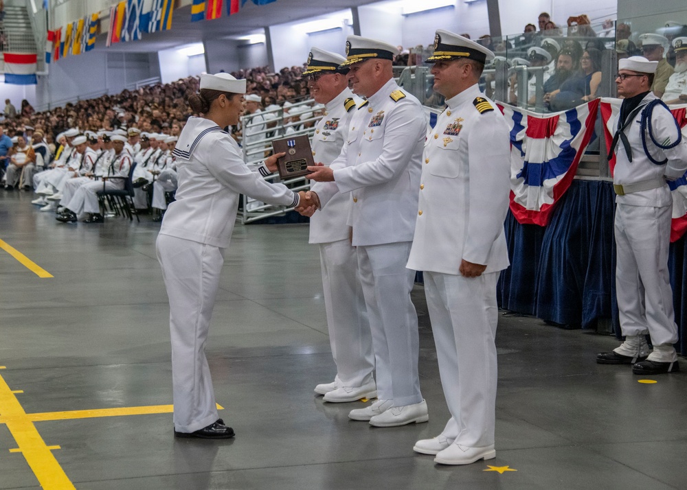 A U.S. Navy military ceremony is taking place in a large indoor venue, likely a graduation or awards ceremony. A female sailor in dress whites is receiving a plaque or award from senior officers, also in dress white uniforms. A large formation of sailors in dress uniforms is seated in the background as an audience.