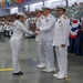A U.S. Navy military ceremony is taking place in a large indoor venue, likely a graduation or awards ceremony. A female sailor in dress whites is receiving a plaque or award from senior officers, also in dress white uniforms. A large formation of sailors in dress uniforms is seated in the background as an audience.