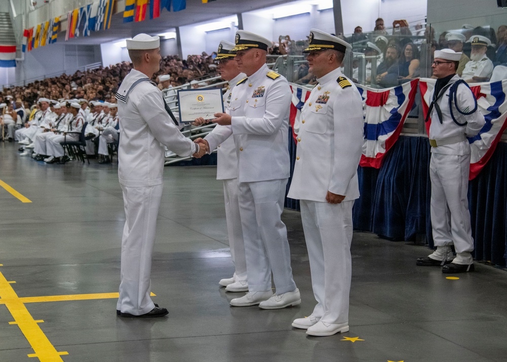 A U.S. Navy military ceremony, likely a graduation or promotion event, is taking place in a large indoor facility. A junior enlisted sailor in dress whites is receiving a certificate while shaking hands with a senior officer, with additional officers and sailors present. A large audience of uniformed personnel and civilians is visible in the background.