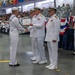 A U.S. Navy military ceremony, likely a graduation or promotion event, is taking place in a large indoor facility. A junior enlisted sailor in dress whites is receiving a certificate while shaking hands with a senior officer, with additional officers and sailors present. A large audience of uniformed personnel and civilians is visible in the background.