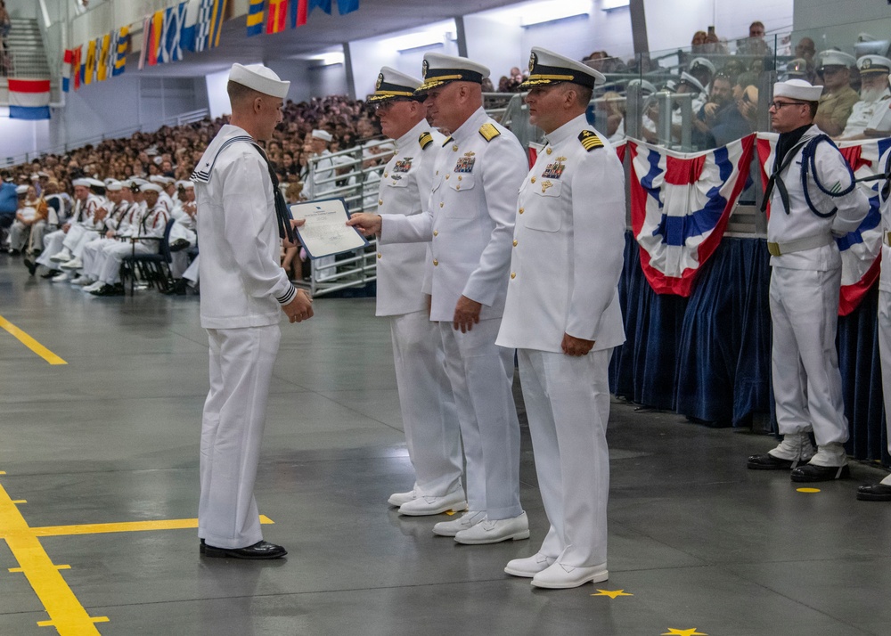 A formal U.S. Navy graduation or awards ceremony taking place in a large indoor venue. A sailor in enlisted dress whites is receiving a certificate or diploma from senior officers in officer dress whites. A large audience of sailors and civilians is seated in the background.