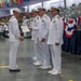 A formal U.S. Navy graduation or awards ceremony taking place in a large indoor venue. A sailor in enlisted dress whites is receiving a certificate or diploma from senior officers in officer dress whites. A large audience of sailors and civilians is seated in the background.