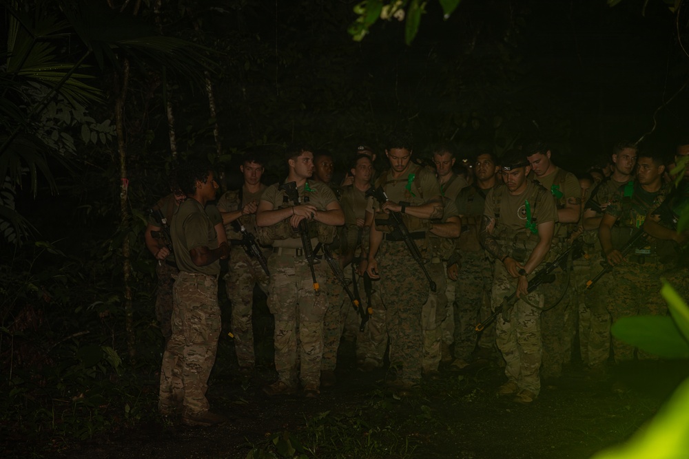 A group of approximately 12-15 armed military personnel are gathered in a dense jungle environment at night. They appear to be receiving a briefing or instructions from one individual on the left side of the group. The personnel are equipped with rifles and wearing camouflage uniforms consistent with a training or operational exercise.