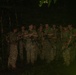 A group of approximately 12-15 armed military personnel are gathered in a dense jungle environment at night. They appear to be receiving a briefing or instructions from one individual on the left side of the group. The personnel are equipped with rifles and wearing camouflage uniforms consistent with a training or operational exercise.