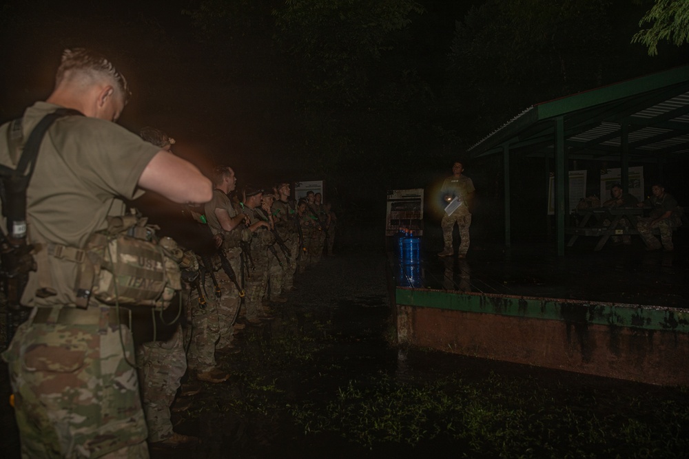 A nighttime military briefing or formation is taking place outdoors, with a group of soldiers in camouflage uniforms lined up facing an instructor or leader standing on an elevated platform. The scene is dimly lit, with the briefer holding a light source illuminating informational boards or posters nearby. The soldiers appear to be equipped with tactical gear and weapons.