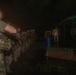 A nighttime military briefing or formation is taking place outdoors, with a group of soldiers in camouflage uniforms lined up facing an instructor or leader standing on an elevated platform. The scene is dimly lit, with the briefer holding a light source illuminating informational boards or posters nearby. The soldiers appear to be equipped with tactical gear and weapons.