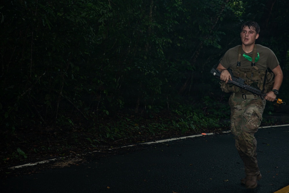 A soldier in military camouflage (OCP/MultiCam pattern) is running along a dark paved road at night or in low-light conditions, carrying what appears to be an M4/M16-style rifle. The individual is wearing a tactical vest, olive drab t-shirt, and has a green bandana or cloth around their neck. The setting appears to be a jungle or heavily forested environment.