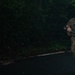 A soldier in military camouflage (OCP/MultiCam pattern) is running along a dark paved road at night or in low-light conditions, carrying what appears to be an M4/M16-style rifle. The individual is wearing a tactical vest, olive drab t-shirt, and has a green bandana or cloth around their neck. The setting appears to be a jungle or heavily forested environment.
