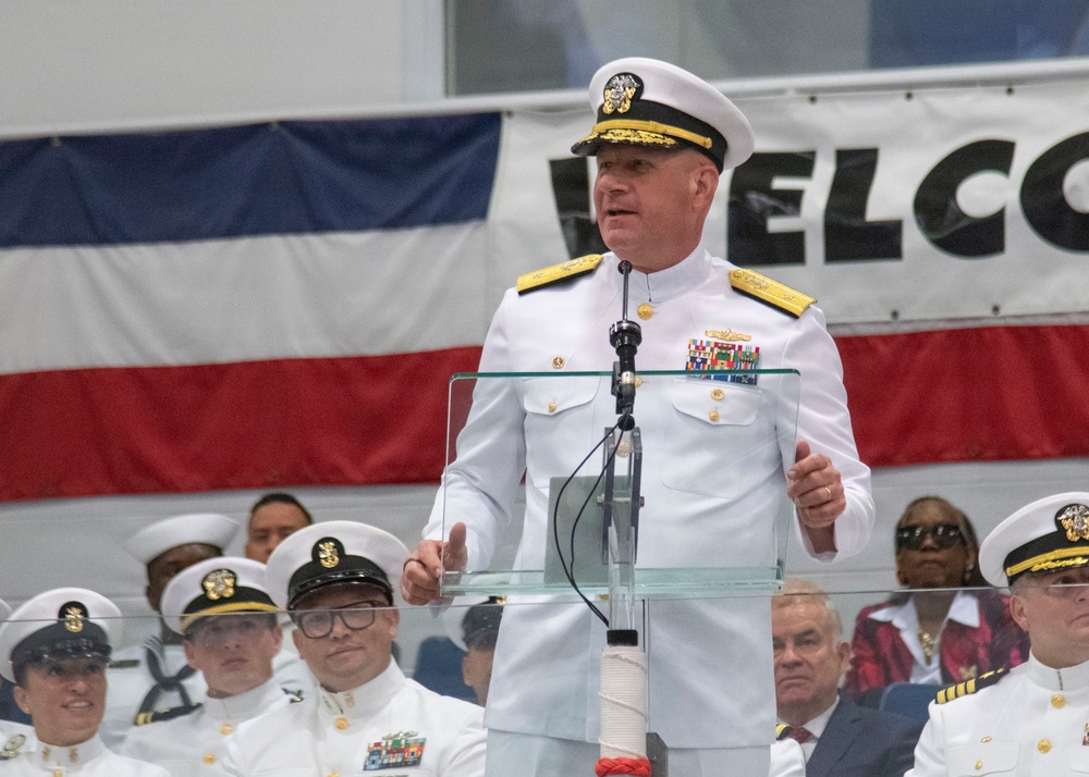 A high-ranking U.S. Navy admiral in dress white uniform is delivering a speech at a formal military ceremony, standing at a clear acrylic podium with a microphone. Behind him, rows of Navy personnel in white uniforms and combination covers are seated as an audience. A partial 'WELCOME' banner and an American flag are visible in the background.