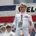 A high-ranking U.S. Navy admiral in dress white uniform is delivering a speech at a formal military ceremony, standing at a clear acrylic podium with a microphone. Behind him, rows of Navy personnel in white uniforms and combination covers are seated as an audience. A partial 'WELCOME' banner and an American flag are visible in the background.