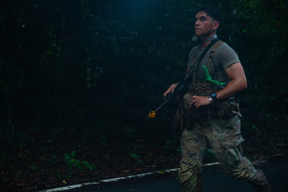 A soldier in military camouflage (MultiCam/OCP pattern) is moving along a paved road at night or in low-light conditions surrounded by dense vegetation. He is carrying a rifle fitted with a yellow training/blank-firing adapter, indicating this is a training exercise rather than a combat situation. He appears alert, scanning his surroundings while moving.