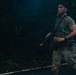 A soldier in military camouflage (MultiCam/OCP pattern) is moving along a paved road at night or in low-light conditions surrounded by dense vegetation. He is carrying a rifle fitted with a yellow training/blank-firing adapter, indicating this is a training exercise rather than a combat situation. He appears alert, scanning his surroundings while moving.