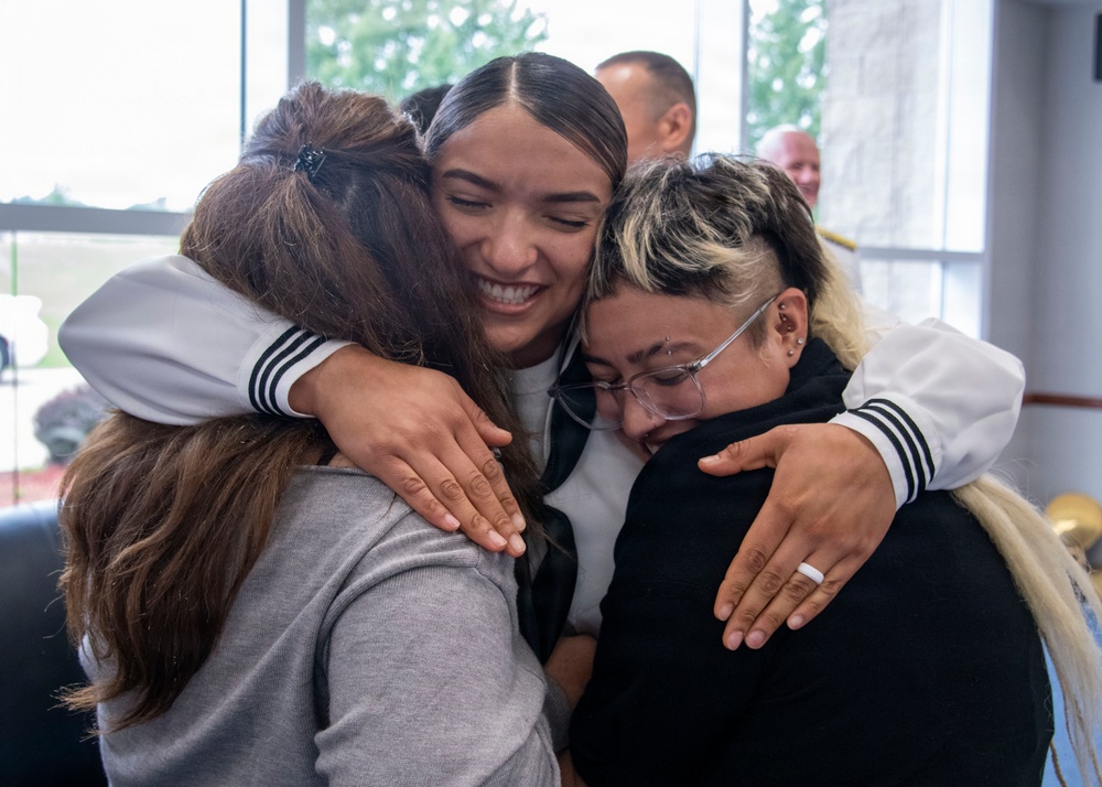 Three young women are sharing a warm group hug, with the central figure smiling broadly. The moment appears emotionally significant, as one of the women on the right appears to be crying or deeply moved. The setting appears to be an indoor facility with windows visible in the background.