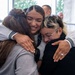 Three young women are sharing a warm group hug, with the central figure smiling broadly. The moment appears emotionally significant, as one of the women on the right appears to be crying or deeply moved. The setting appears to be an indoor facility with windows visible in the background.