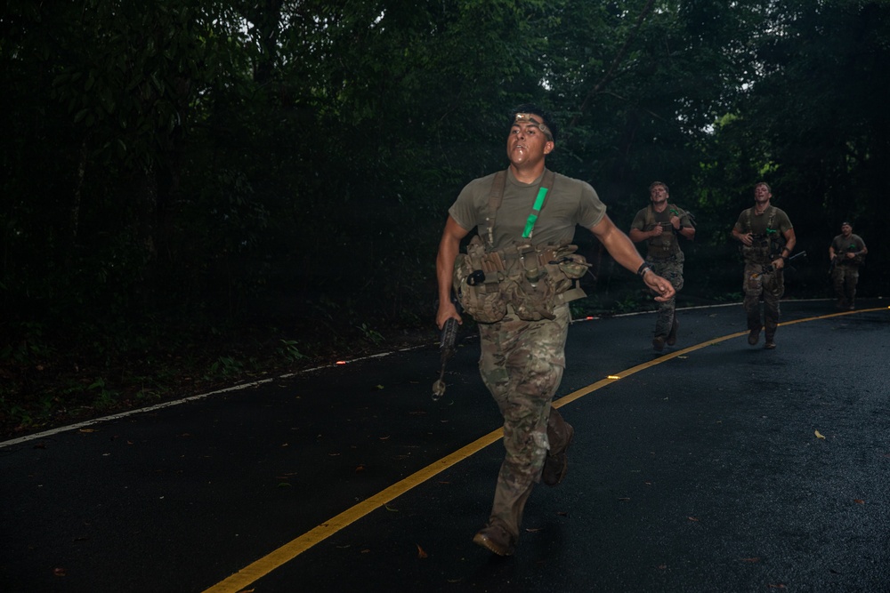A group of military personnel in combat gear are running along a wet, curved asphalt road bordered by dense tropical vegetation. The lead soldier is wearing multicam fatigues, a tactical vest, and carries a weapon, with a green glow stick attached to his gear. The image appears to capture a nighttime or low-light training exercise or tactical movement.