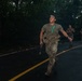 A group of military personnel in combat gear are running along a wet, curved asphalt road bordered by dense tropical vegetation. The lead soldier is wearing multicam fatigues, a tactical vest, and carries a weapon, with a green glow stick attached to his gear. The image appears to capture a nighttime or low-light training exercise or tactical movement.