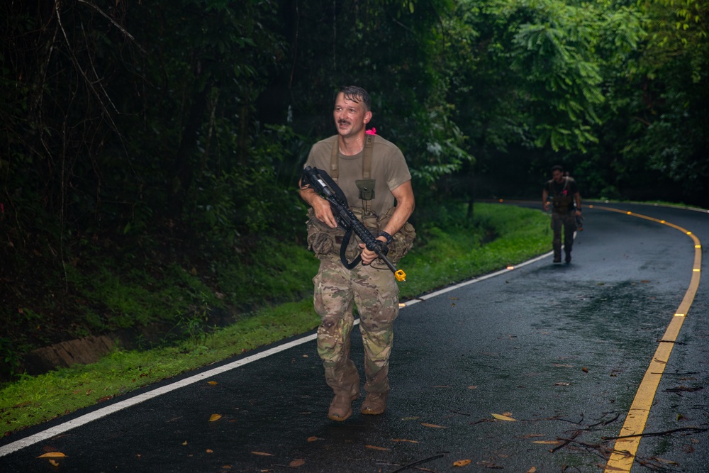 Two individuals in military-style camouflage uniforms are moving along a wet, curved road surrounded by dense tropical vegetation. The foreground figure is carrying a rifle and wearing tactical gear, appearing wet as if in rainy conditions. A second similarly equipped individual is visible further down the road in the background.