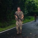 Two individuals in military-style camouflage uniforms are moving along a wet, curved road surrounded by dense tropical vegetation. The foreground figure is carrying a rifle and wearing tactical gear, appearing wet as if in rainy conditions. A second similarly equipped individual is visible further down the road in the background.