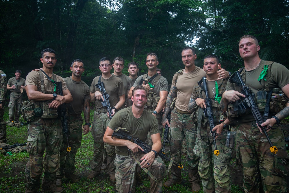A group of approximately 11-12 military personnel pose for a group photo in a jungle or dense forest environment. They are wearing mixed combat uniforms including MultiCam/OCP pattern and olive drab t-shirts, and several are holding rifles with colored training barrel blockers. The setting and equipment suggest a joint military training exercise.