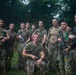 A group of approximately 11-12 military personnel pose for a group photo in a jungle or dense forest environment. They are wearing mixed combat uniforms including MultiCam/OCP pattern and olive drab t-shirts, and several are holding rifles with colored training barrel blockers. The setting and equipment suggest a joint military training exercise.