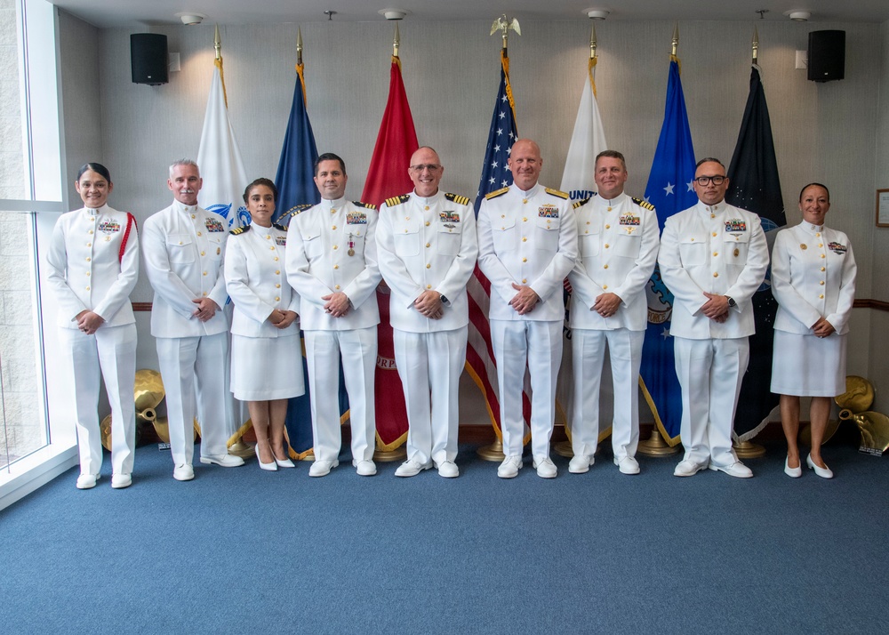 A formal group photograph of nine military personnel dressed in white naval dress uniforms, posing in front of a display of multiple flags including the United States flag. The setting appears to be an official military facility or ceremonial room, likely during a promotion, change of command, or other formal military ceremony. The individuals display various ranks and ribbons/medals on their uniforms.