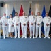 A formal group photograph of nine military personnel dressed in white naval dress uniforms, posing in front of a display of multiple flags including the United States flag. The setting appears to be an official military facility or ceremonial room, likely during a promotion, change of command, or other formal military ceremony. The individuals display various ranks and ribbons/medals on their uniforms.