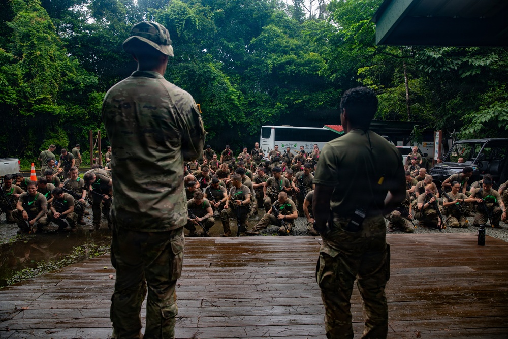 Two military instructors or leaders stand on an elevated wooden platform addressing a large group of soldiers crouched on wet gravel ground. The setting appears to be a jungle or tropical training environment, with dense green foliage in the background. The soldiers are wearing camouflage uniforms with green markers/patches, suggesting a training exercise or briefing.