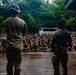 Two military instructors or leaders stand on an elevated wooden platform addressing a large group of soldiers crouched on wet gravel ground. The setting appears to be a jungle or tropical training environment, with dense green foliage in the background. The soldiers are wearing camouflage uniforms with green markers/patches, suggesting a training exercise or briefing.