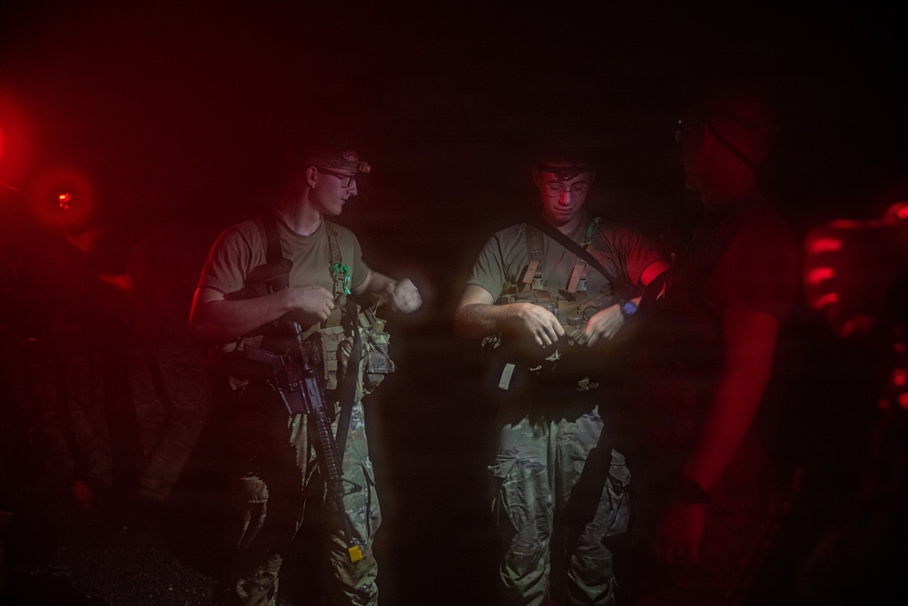 Two armed military or tactical personnel are visible in a dark, red-lit environment, appearing to conduct equipment checks or gear adjustments. The scene is illuminated by a combination of red ambient light and a white light source, suggesting a nighttime operational setting. Additional personnel are partially visible in the background.