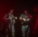 Two armed military or tactical personnel are visible in a dark, red-lit environment, appearing to conduct equipment checks or gear adjustments. The scene is illuminated by a combination of red ambient light and a white light source, suggesting a nighttime operational setting. Additional personnel are partially visible in the background.