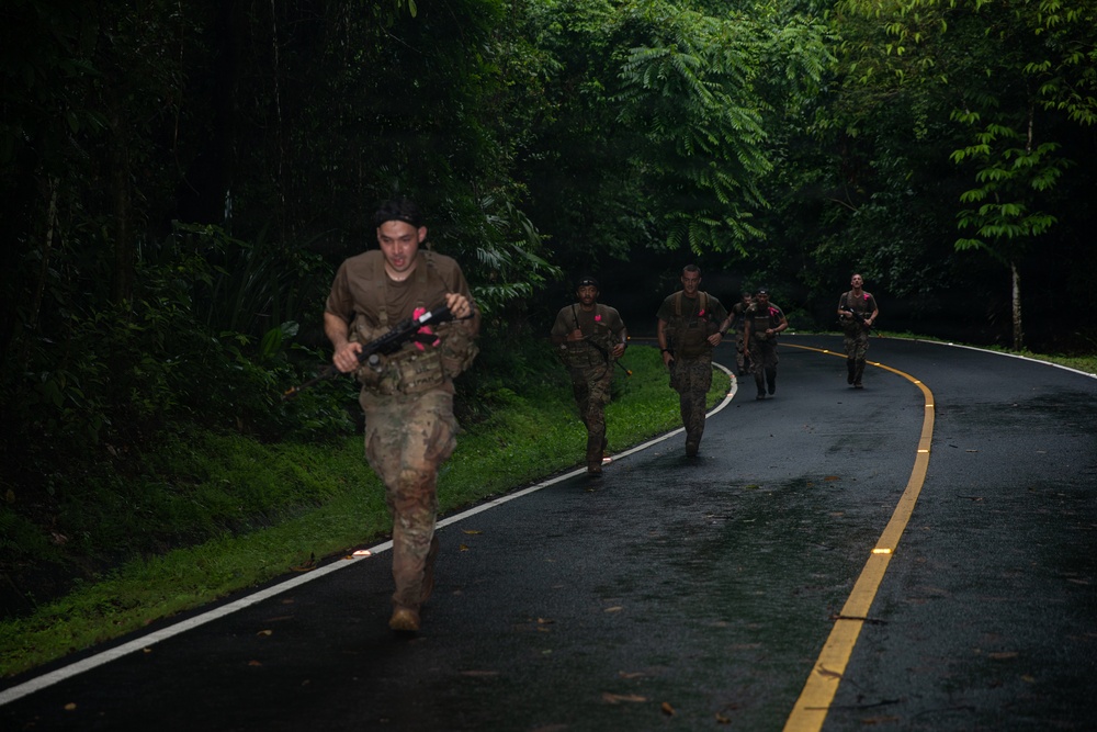 A group of military personnel in camouflage uniforms are running along a wet, paved road flanked by dense tropical jungle vegetation. The lead soldier is carrying a weapon with pink/red markings, suggesting training or exercise ammunition. The scene appears to be a military physical training or tactical exercise conducted in a tropical environment.
