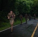 A group of military personnel in camouflage uniforms are running along a wet, paved road flanked by dense tropical jungle vegetation. The lead soldier is carrying a weapon with pink/red markings, suggesting training or exercise ammunition. The scene appears to be a military physical training or tactical exercise conducted in a tropical environment.