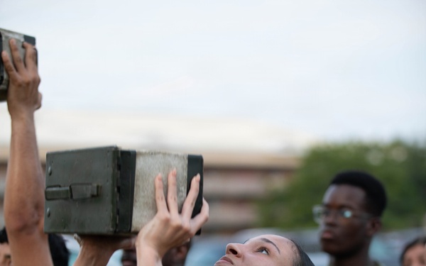A female military service member in U.S. Marine Corps utility uniform is lifting a military ammunition can overhead during what appears to be a physical fitness training exercise. She is wearing a pack harness and a high-visibility reflective belt, standard PT safety gear. Other uniformed personnel are visible in the background observing or participating.
